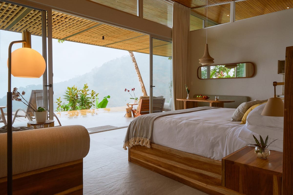 A modern bedroom with a large bed, wooden headboard, and a view of mountains through large windows.