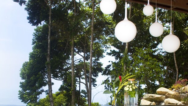Un espace aménagé au bord de la piscine, avec une terrasse en bois, un bar doté de tabourets, des luminaires sphériques suspendus et une vue sur l’océan et les arbres.