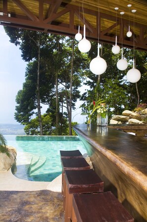 Zona de piscina con terraza de madera, bar con taburetes, lámparas esféricas colgantes y vistas al océano y árboles.