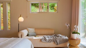 A bedroom with a bed, bedside table, lamp, and a window with a view of greenery.