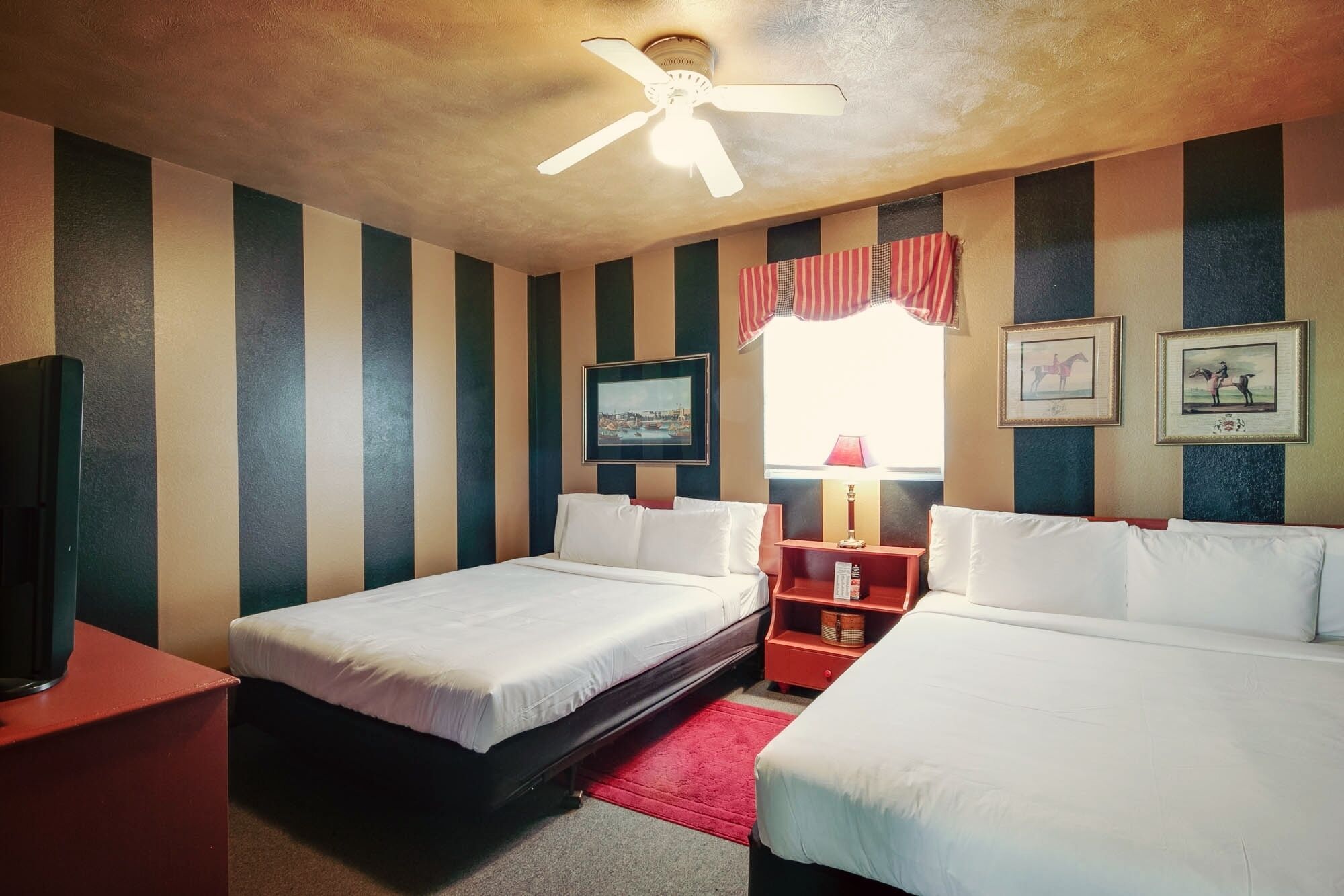 A hotel room with two beds, a ceiling fan, a window with curtains, and framed pictures on the wall.
