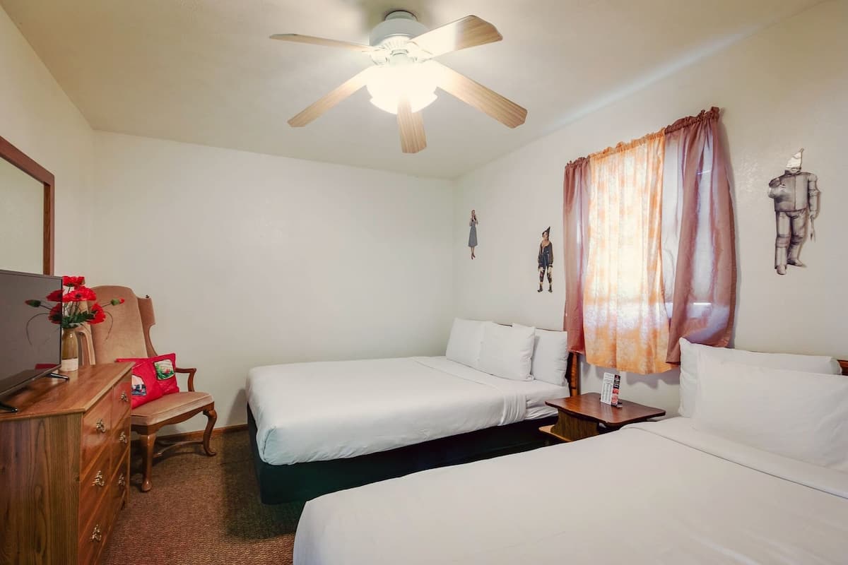 Quarto de hotel com duas camas, uma escrivaninha de madeira com cadeira, ventilador de teto e uma parede com obras de arte.