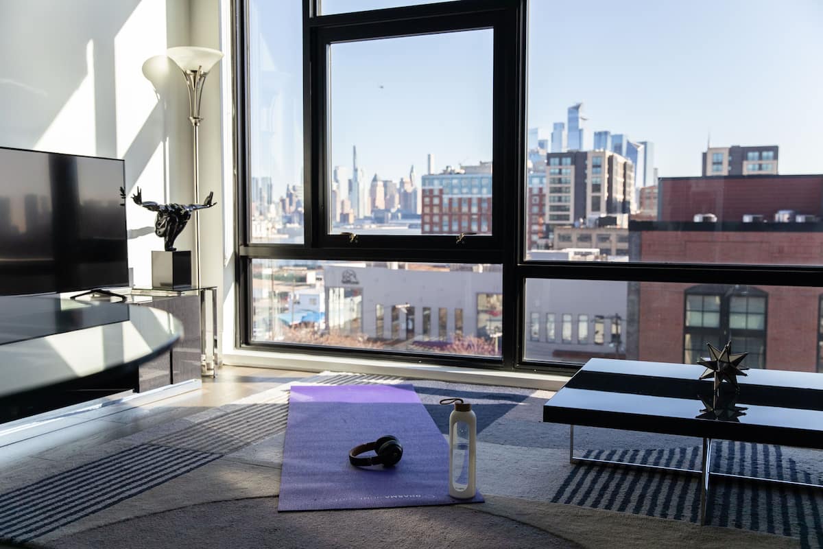 Una habitación moderna con un gran ventanal, una esterilla de yoga, auriculares, una botella de agua y un escritorio minimalista.