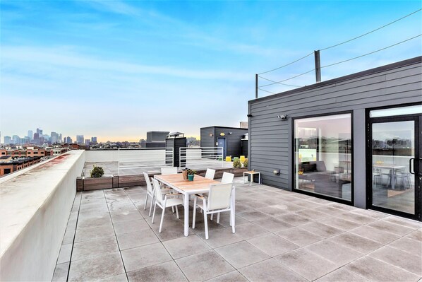A rooftop patio with a dining table and chairs, a small garden area, and a city skyline view.