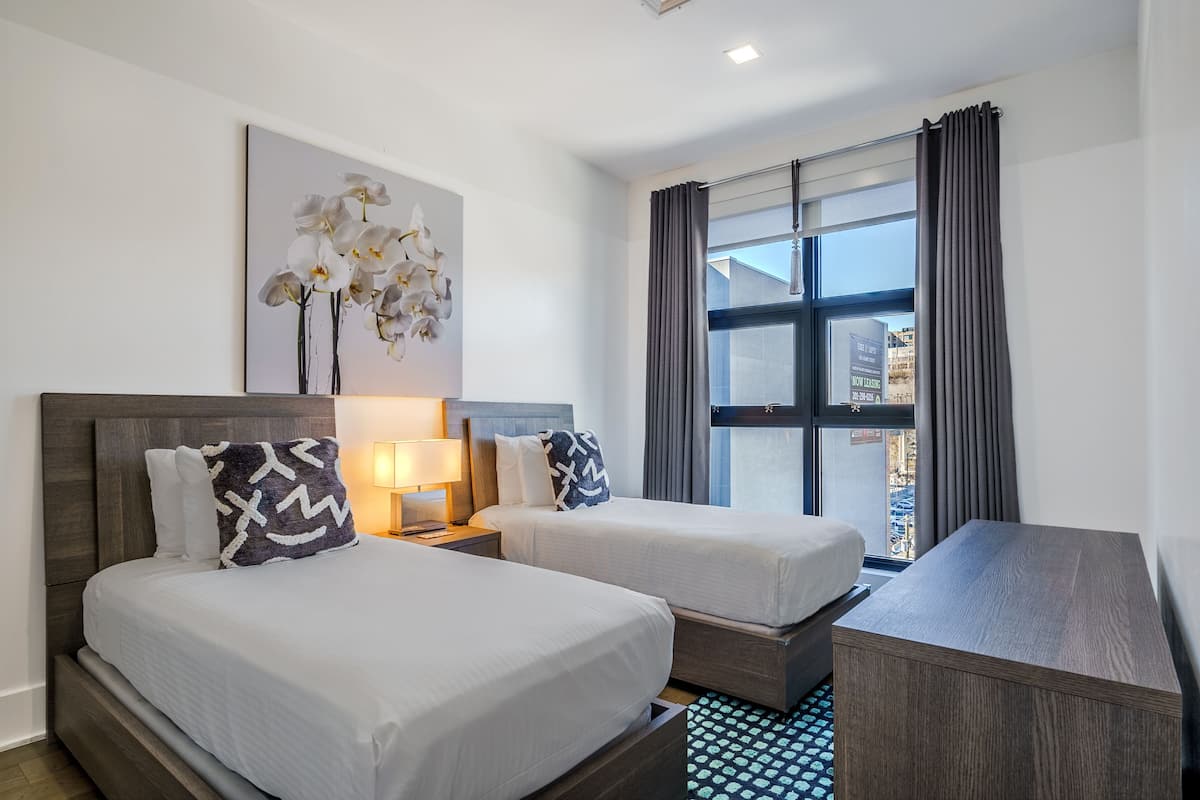 Una habitación de hotel con dos camas, un escritorio, una lámpara, un ventanal grande con cortinas y una obra de arte en la pared de orquídeas blancas.