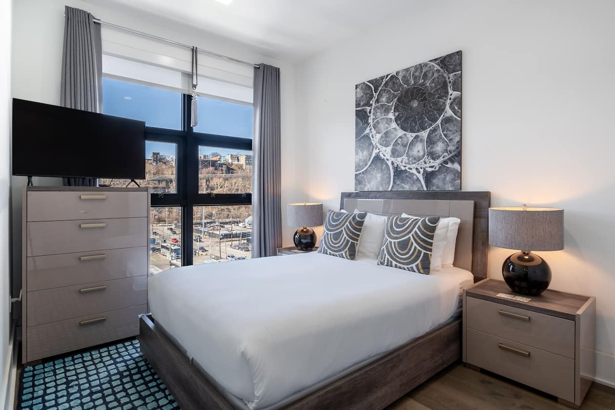 Una habitación de hotel moderna con una cama grande, un televisor de pantalla plana, una mesita de noche con una lámpara y vistas a la ciudad a través de la ventana.