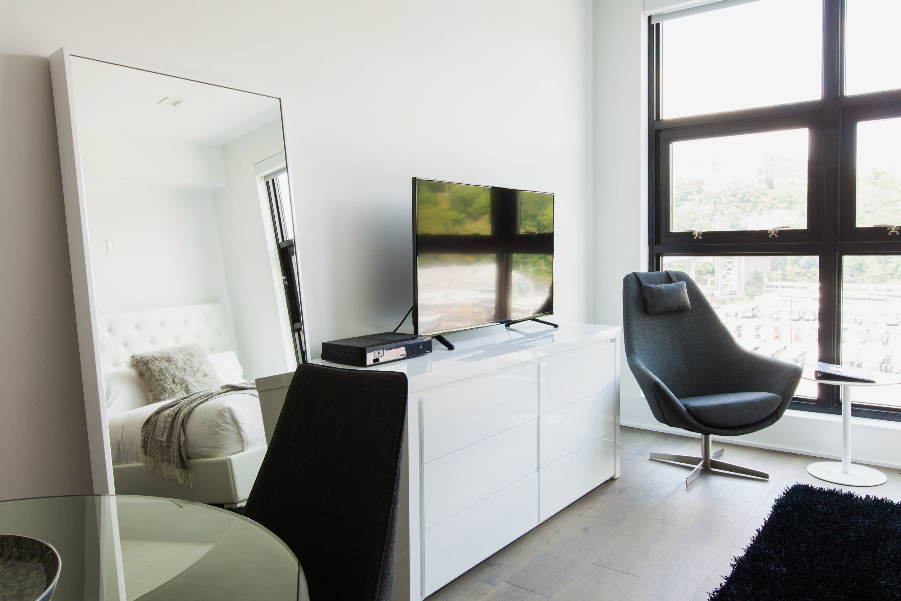 A modern living room with a white sofa, a black armchair, a glass coffee table, a flat-screen TV on a white cabinet, and a large window with a view of buildings.