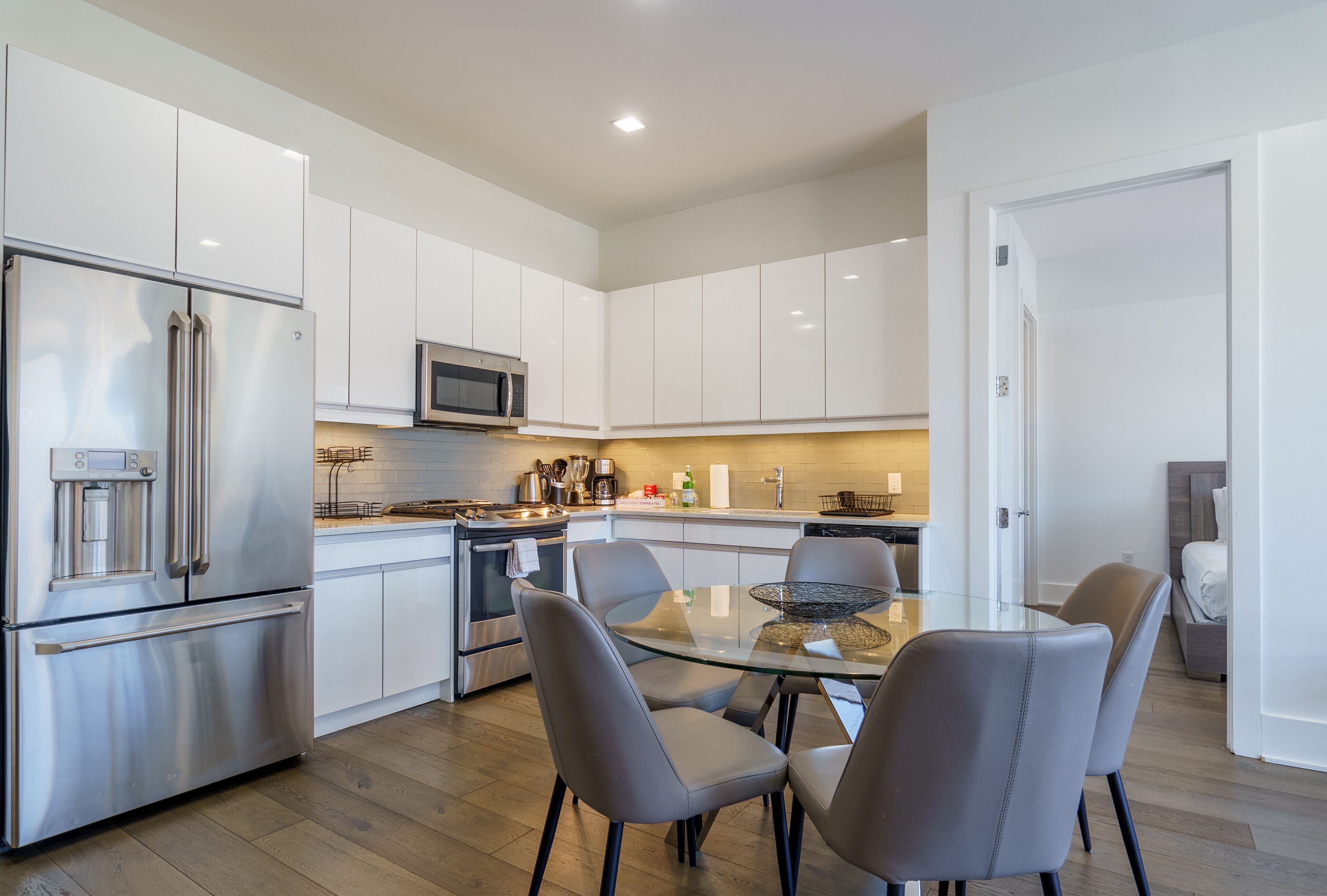 A modern kitchen with white cabinets, stainless steel appliances, and a glass dining table with four chairs.