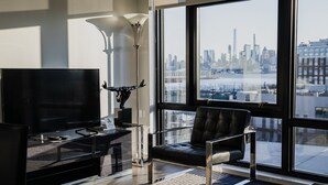 Un salon moderne doté d’un grand téléviseur, d’un fauteuil en cuir noir, d’un lampadaire et offrant une vue sur les gratte-ciel de la ville à travers de grandes fenêtres.