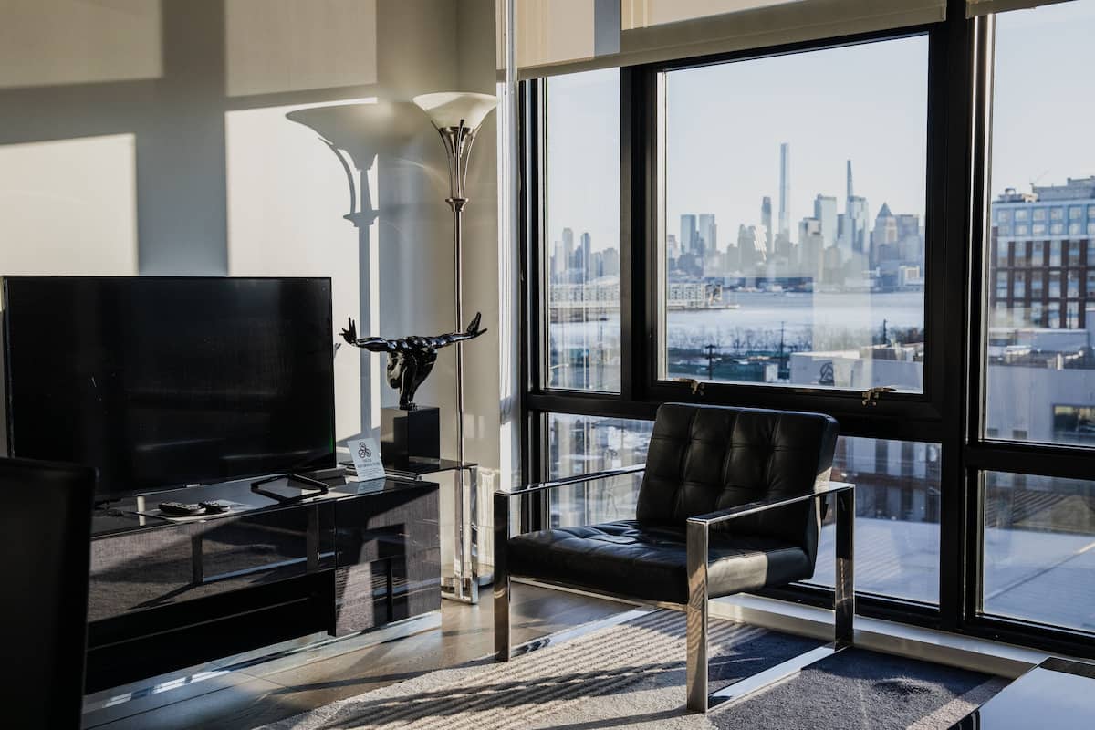 Una sala de estar moderna con un televisor grande, un sillón de cuero negro, una lámpara de pie y vistas a un horizonte urbano a través de ventanas amplias.