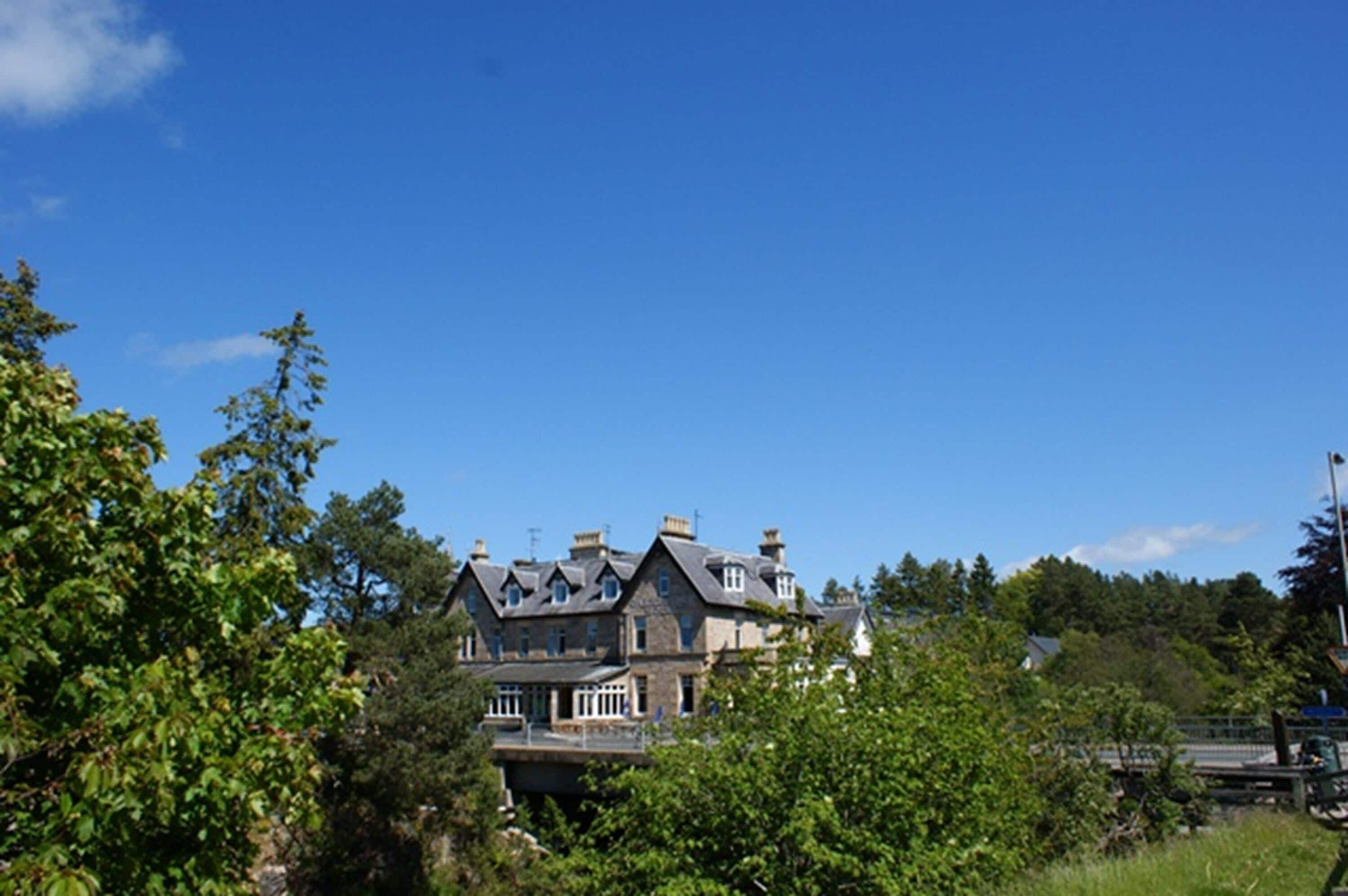Un edificio grande y antiguo de piedra con varias chimeneas, rodeado de abundante vegetación y árboles.