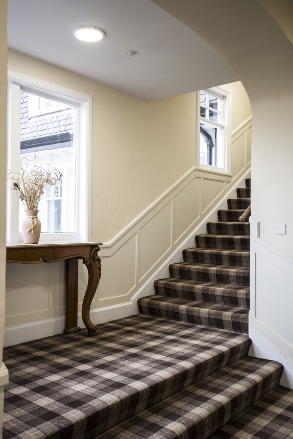 A staircase with a checkered carpet, a wooden console table with a vase, and a window with a view of a building. - Carrbridge Hotel (Carrbridge)
