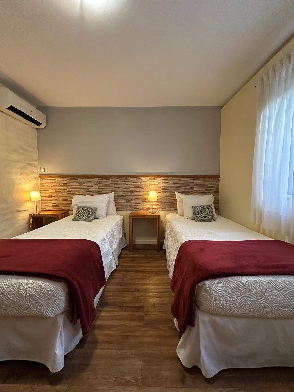 A room with two beds, wooden headboard, bedside lamps, and a window with curtains.