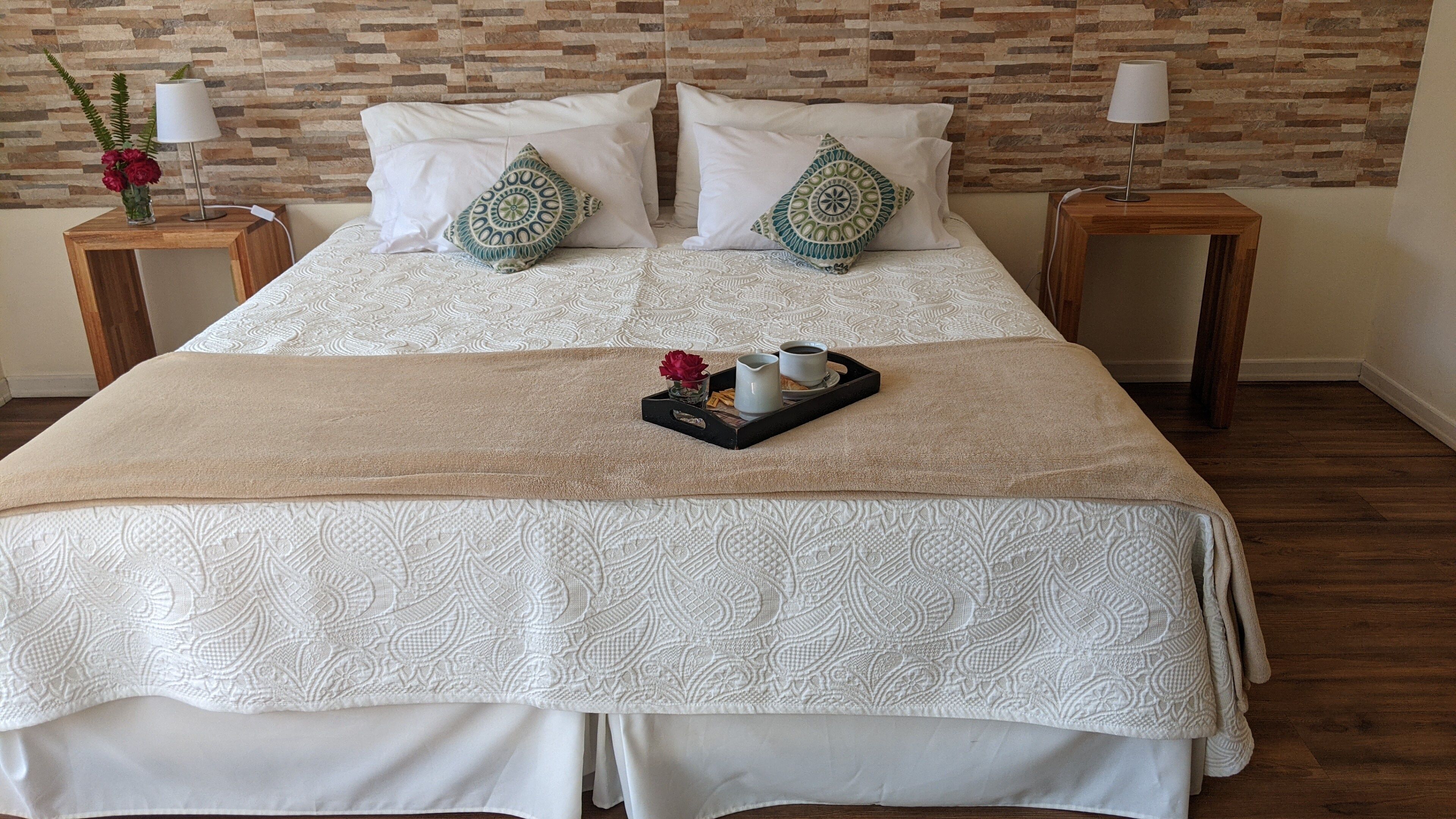 A neatly made bed with a patterned bedspread, two pillows, a tray with a cup and a small vase of flowers, and a nightstand with a lamp.