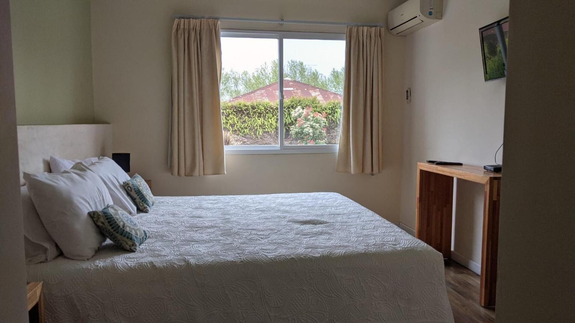 Een slaapkamer met een bed, gordijnen, een airconditioning en een raam met uitzicht op groen en een gebouw.
