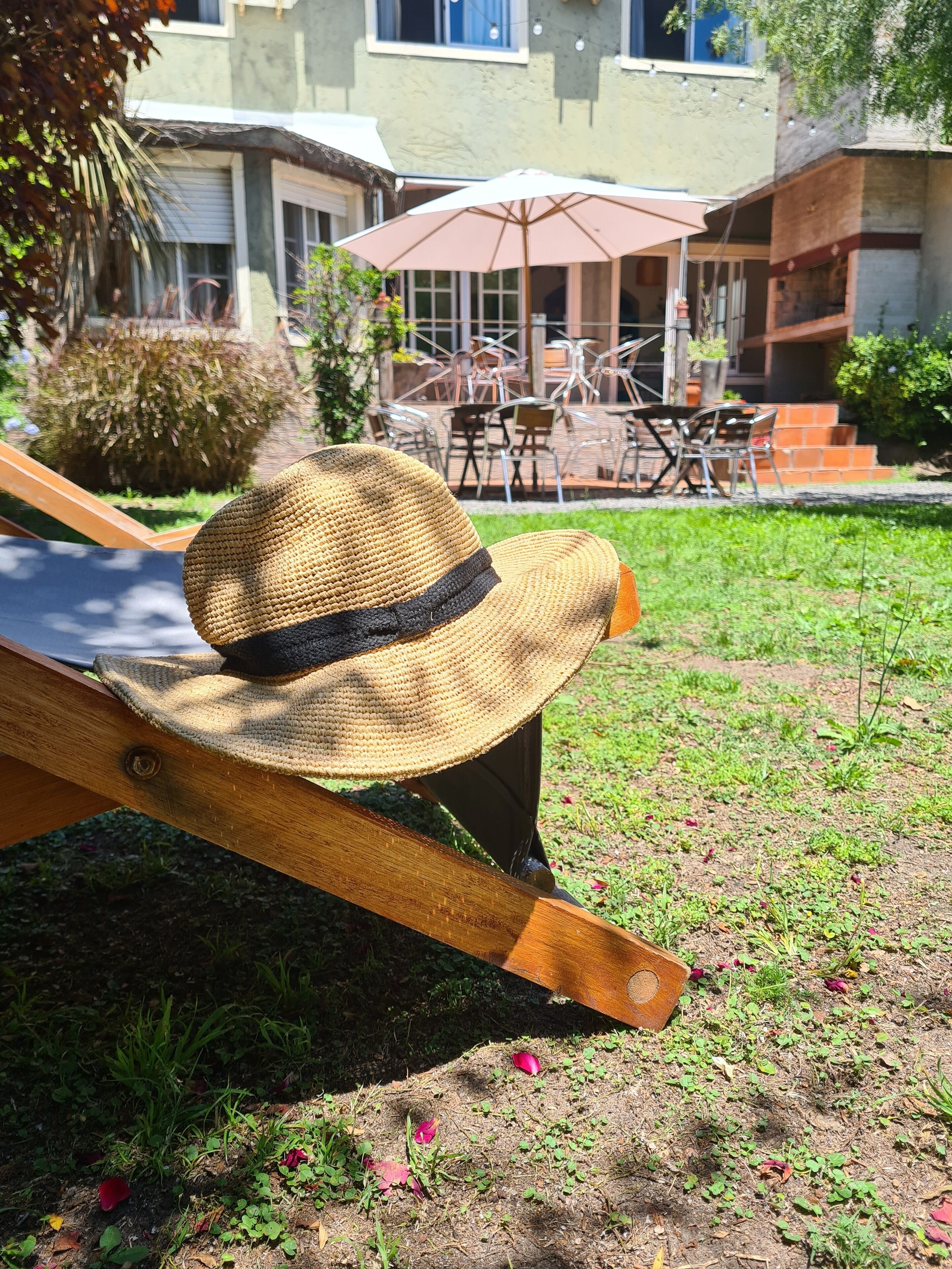 A straw hat on a wooden lounge chair in a garden area with outdoor furniture and a building in the background.