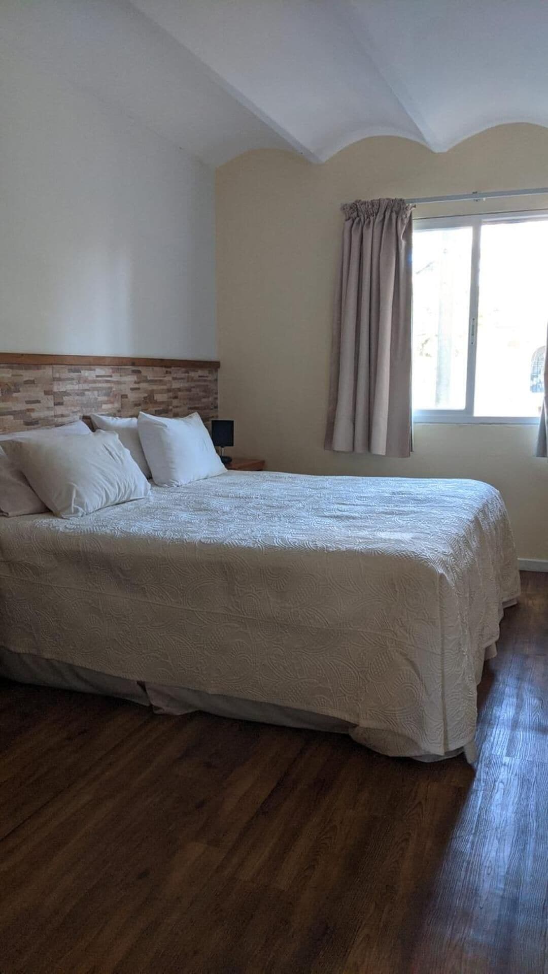 A bedroom with a large bed, a wooden headboard, a nightstand, a window with curtains, and a brick-patterned wall.