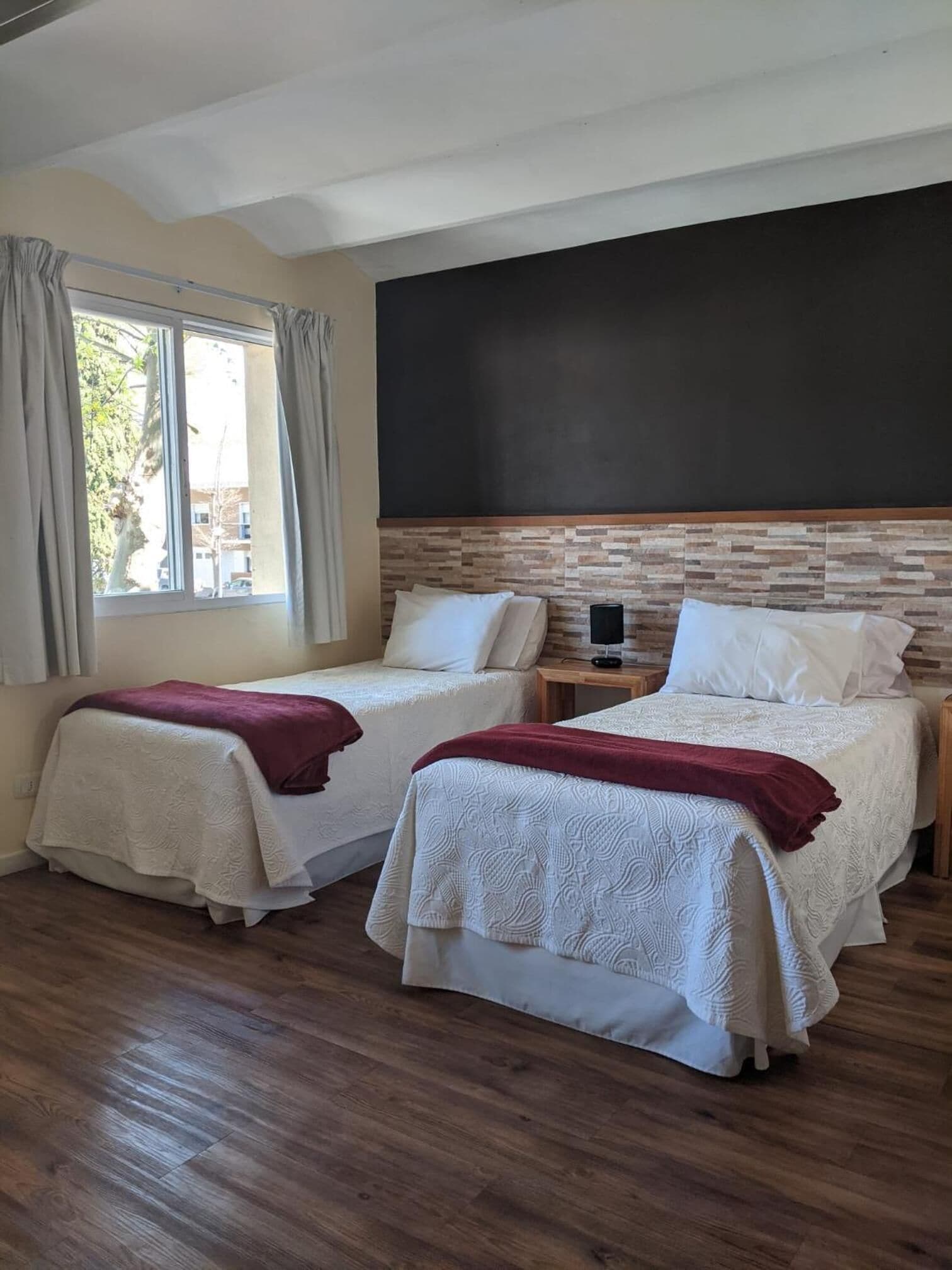 Ein Zimmer mit zwei Betten, einem großen Fenster mit Vorhängen, einem Holzboden und einer Tafel an der Wand.