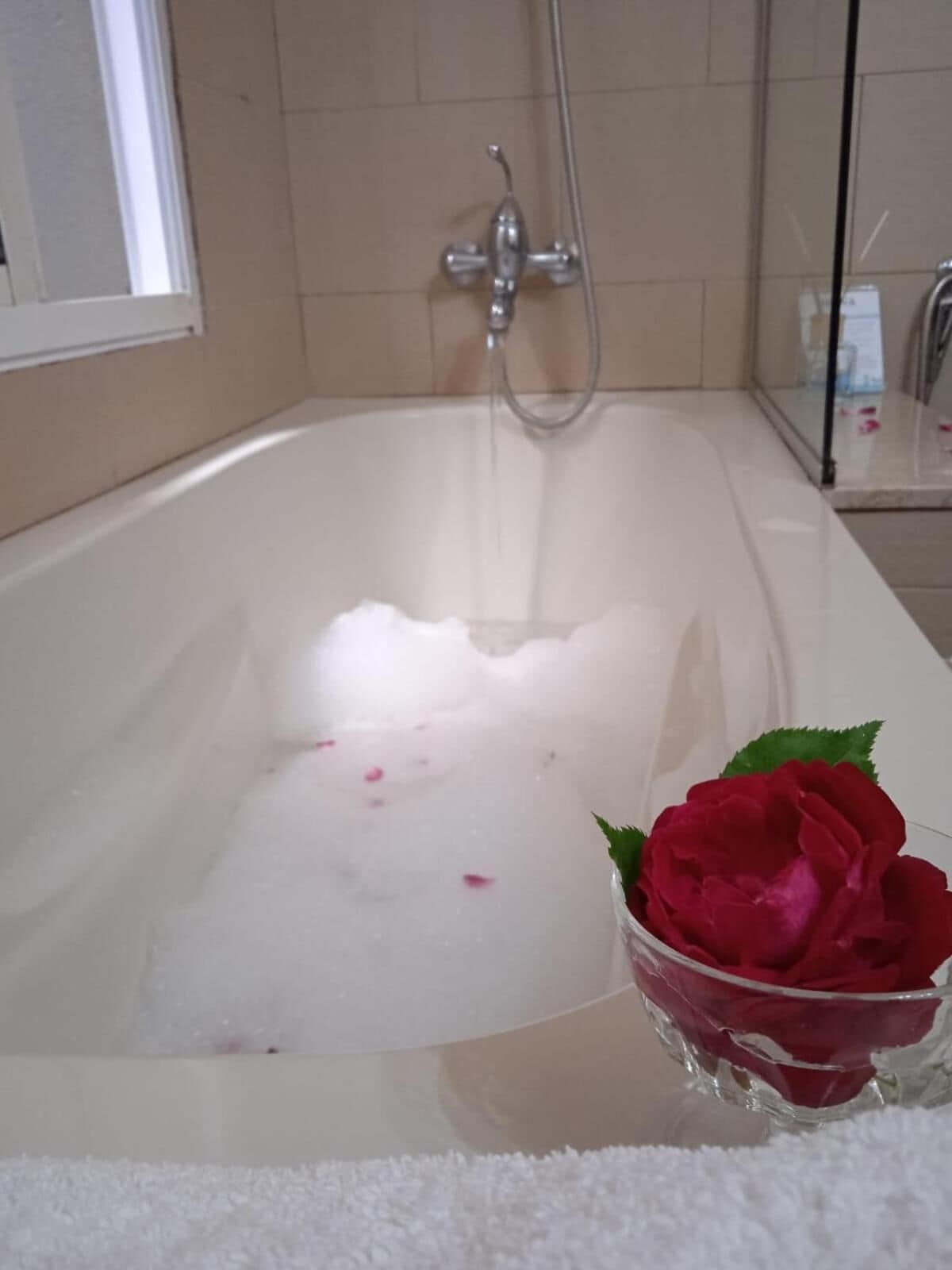 Una bañera con agua y pétalos de rosa, un cuenco de vidrio con una rosa roja, y una toalla blanca en el suelo.