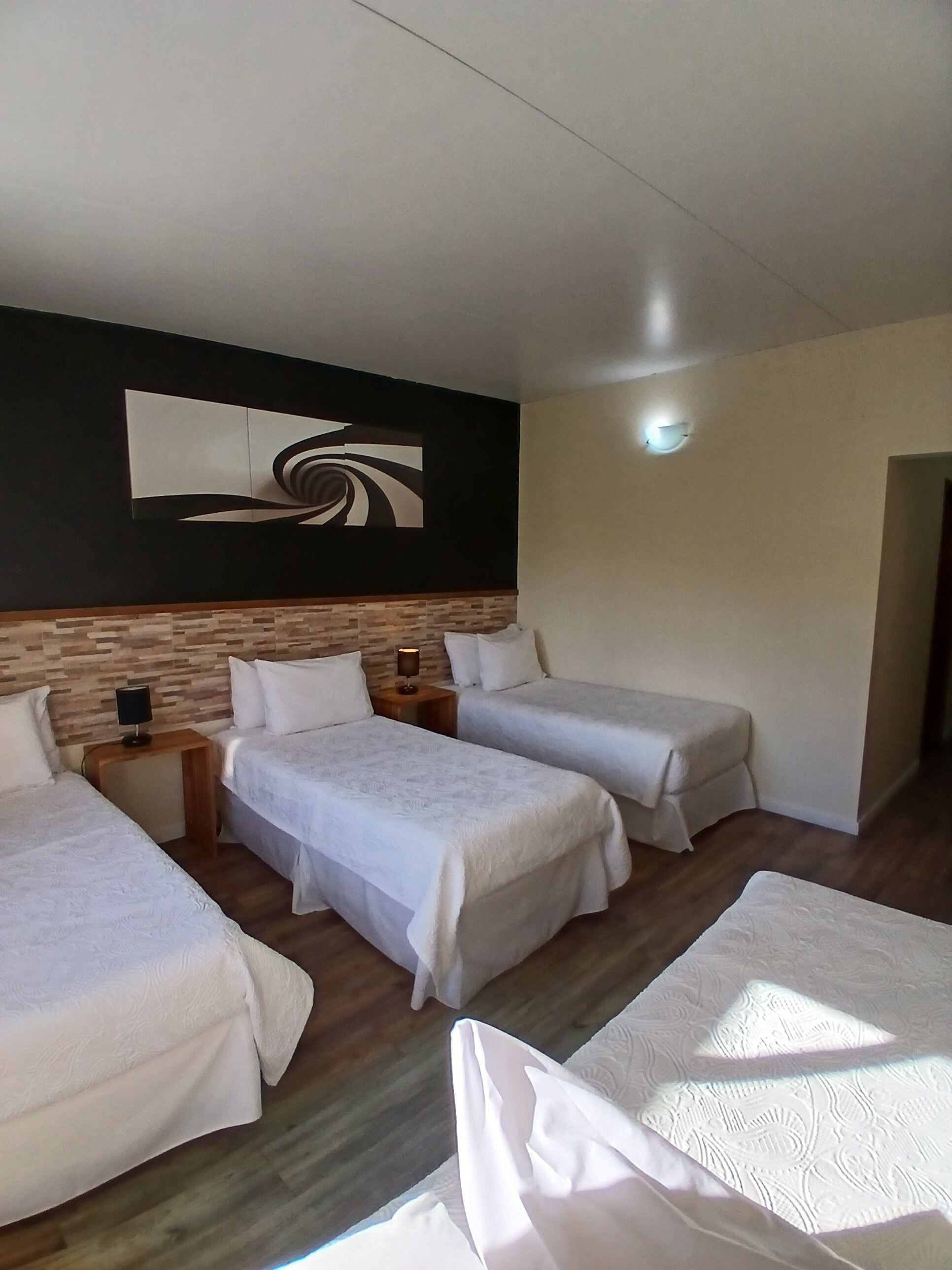 A hotel room with two single beds, a wooden headboard, a wall-mounted artwork, a bedside table with a lamp, and a small bench by the window.