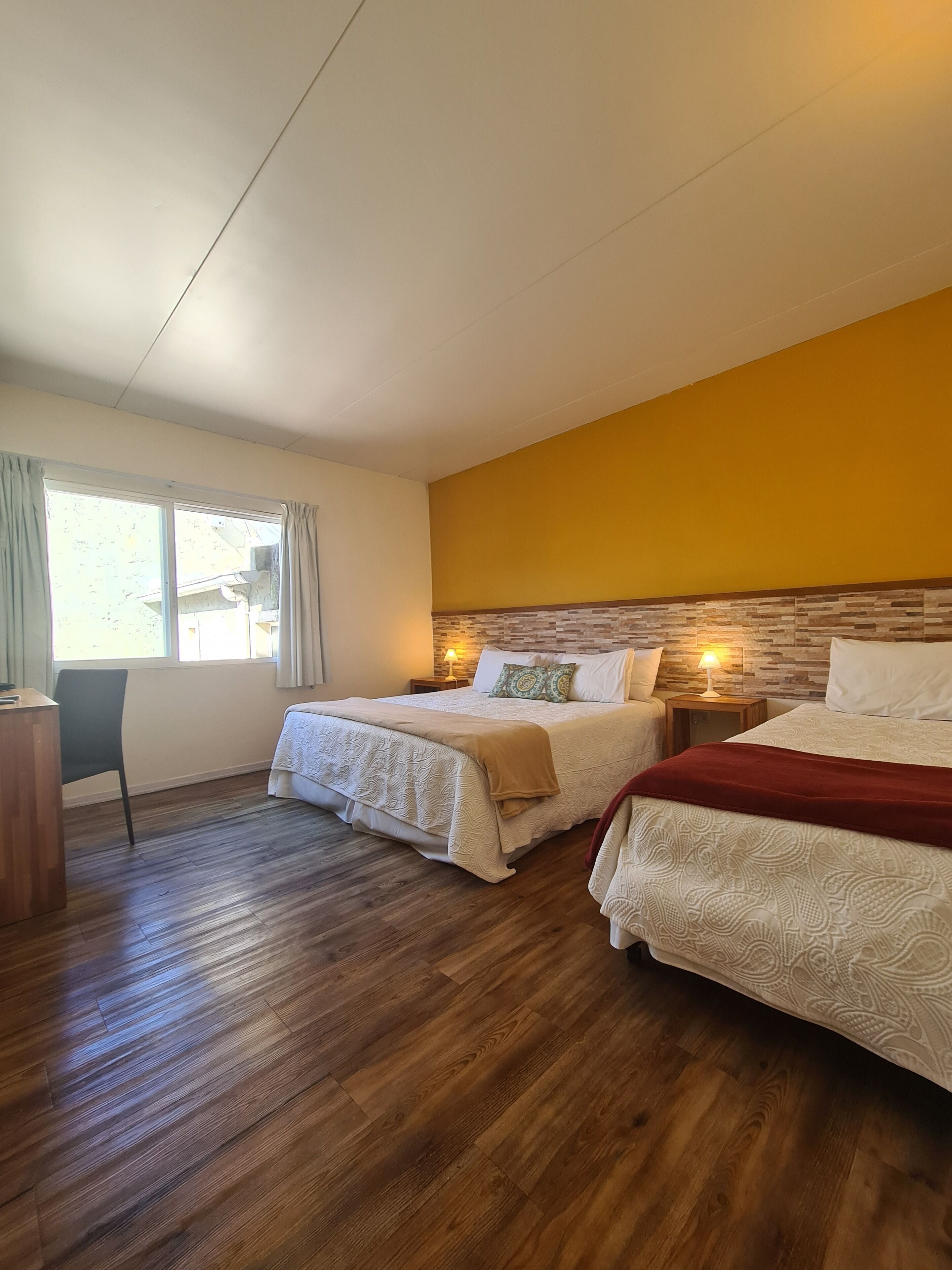 A hotel room with two beds, wooden flooring, a desk, and a window with curtains.