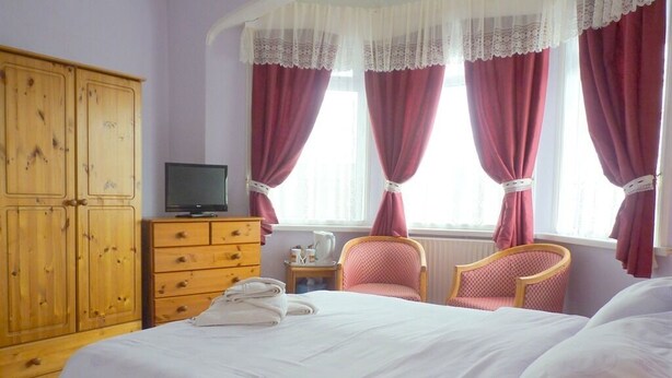A bedroom with a bed, a wooden wardrobe, a television, two chairs, and a window with curtains. - Arendale Hotel (Blackpool)