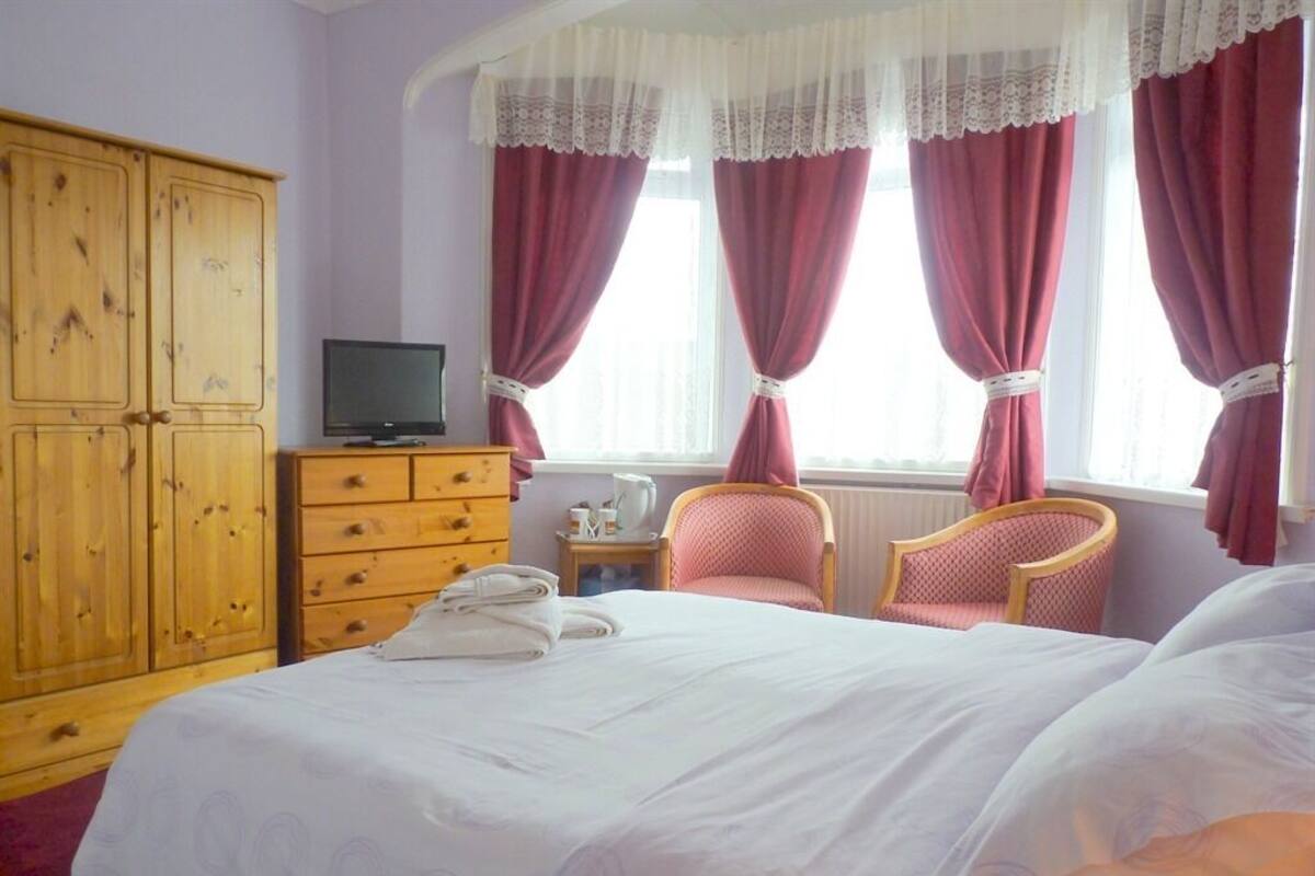 A bedroom with a bed, a wooden wardrobe, a television, two chairs, and a window with curtains.