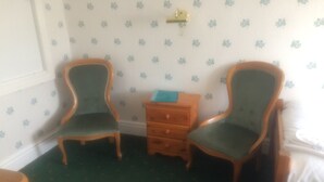 A room with two wooden chairs with green upholstery, a small wooden dresser, and a patterned wallpaper with a floral design. - Arendale Hotel (Blackpool)