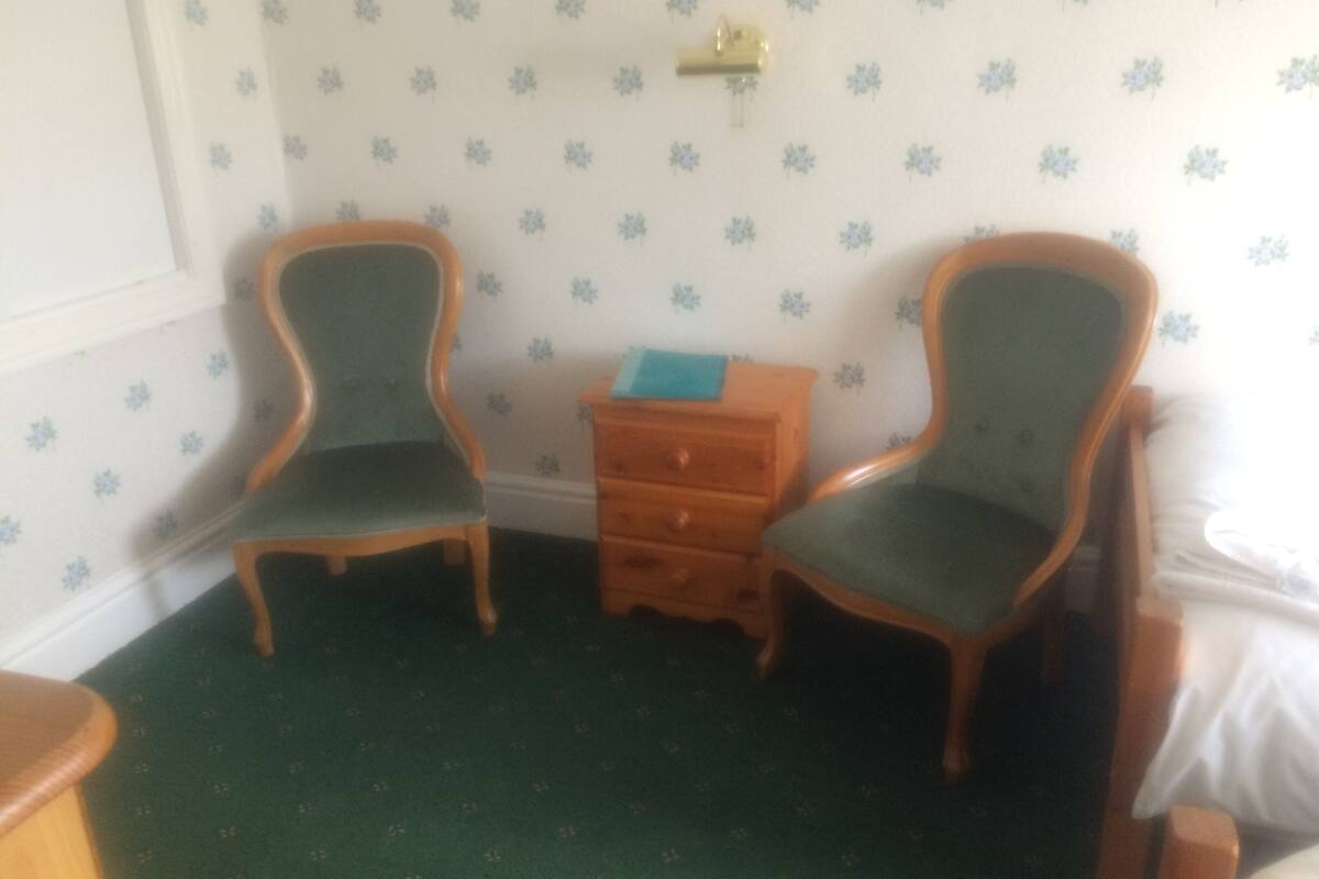 A room with two wooden chairs with green upholstery, a small wooden dresser, and a patterned wallpaper with a floral design.