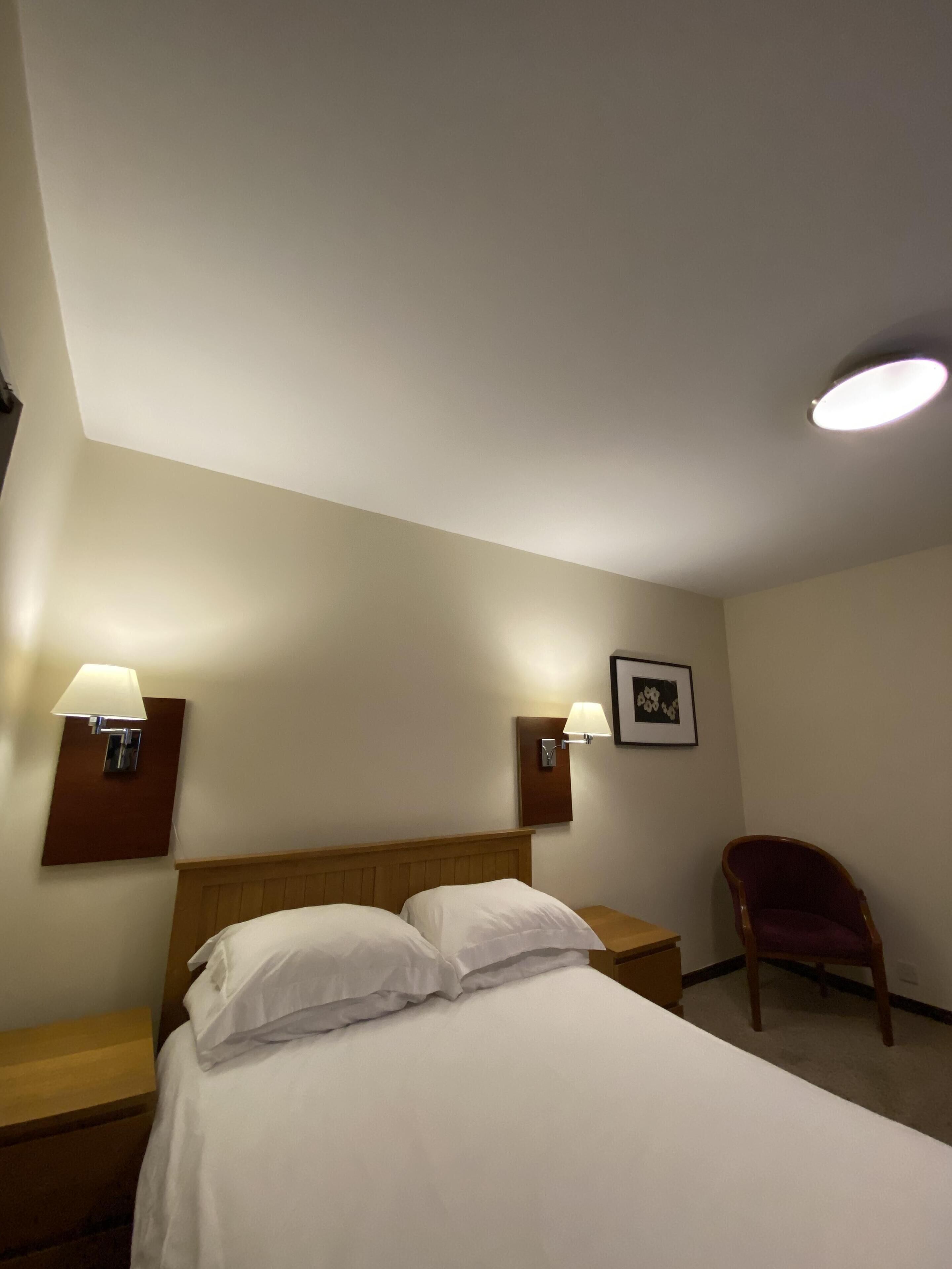 Habitación de hotel con cama, dos mesitas de noche, dos lámparas de pared, un cuadro enmarco y una silla de madera.