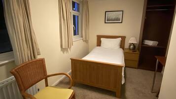 A bedroom with a wooden bed, a chair, a bedside table, a lamp, a window with curtains, and a wardrobe.