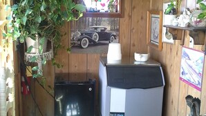 A compact room with a refrigerator, a chair, a table with a basket, and a wall-mounted shelf with decorative items. - Boron Motel (Boron)
