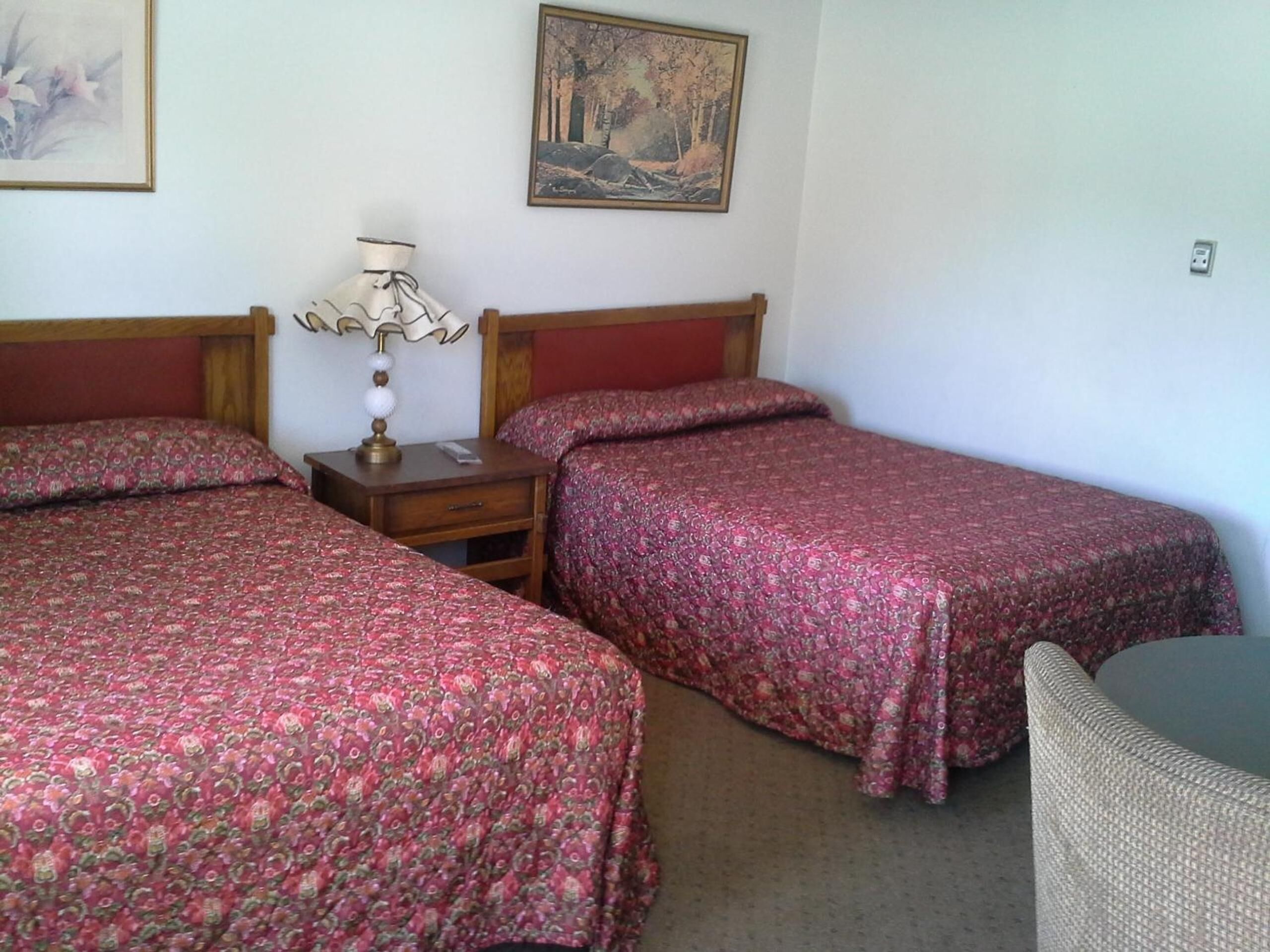 A room with two single beds, a nightstand with a lamp, a chair, and two framed pictures on the wall.