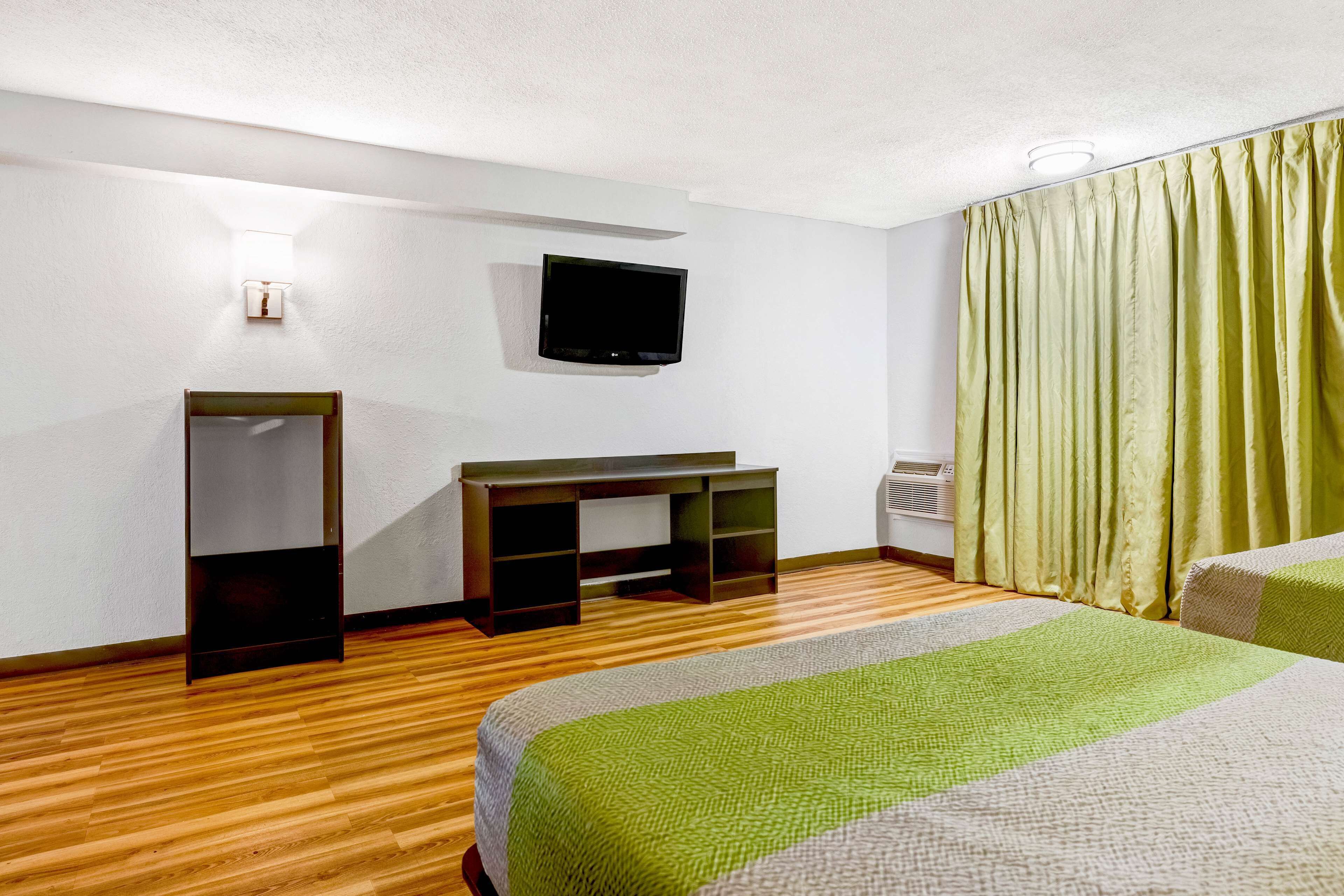A hotel room with a wooden floor, a bed with a green and white bedspread, a desk with a flat-screen TV, a wardrobe, and a window with green curtains.
