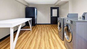 A laundry room with a white table, two washing machines, a refrigerator, and a door. - Motel 6 Indianapolis, IN (Indianapolis)