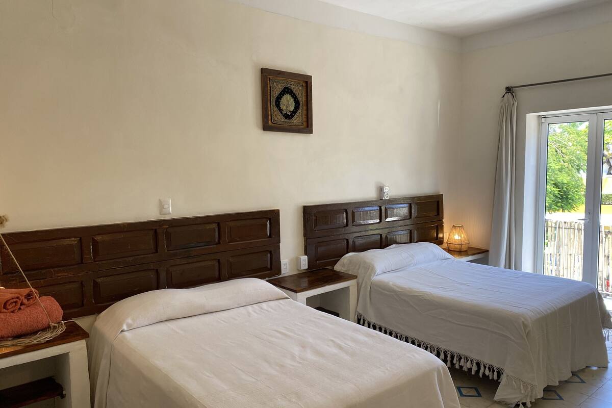 A hotel room with two beds, a wooden headboard, a small table with a towel, a wall-mounted light, and a window with a view of greenery.