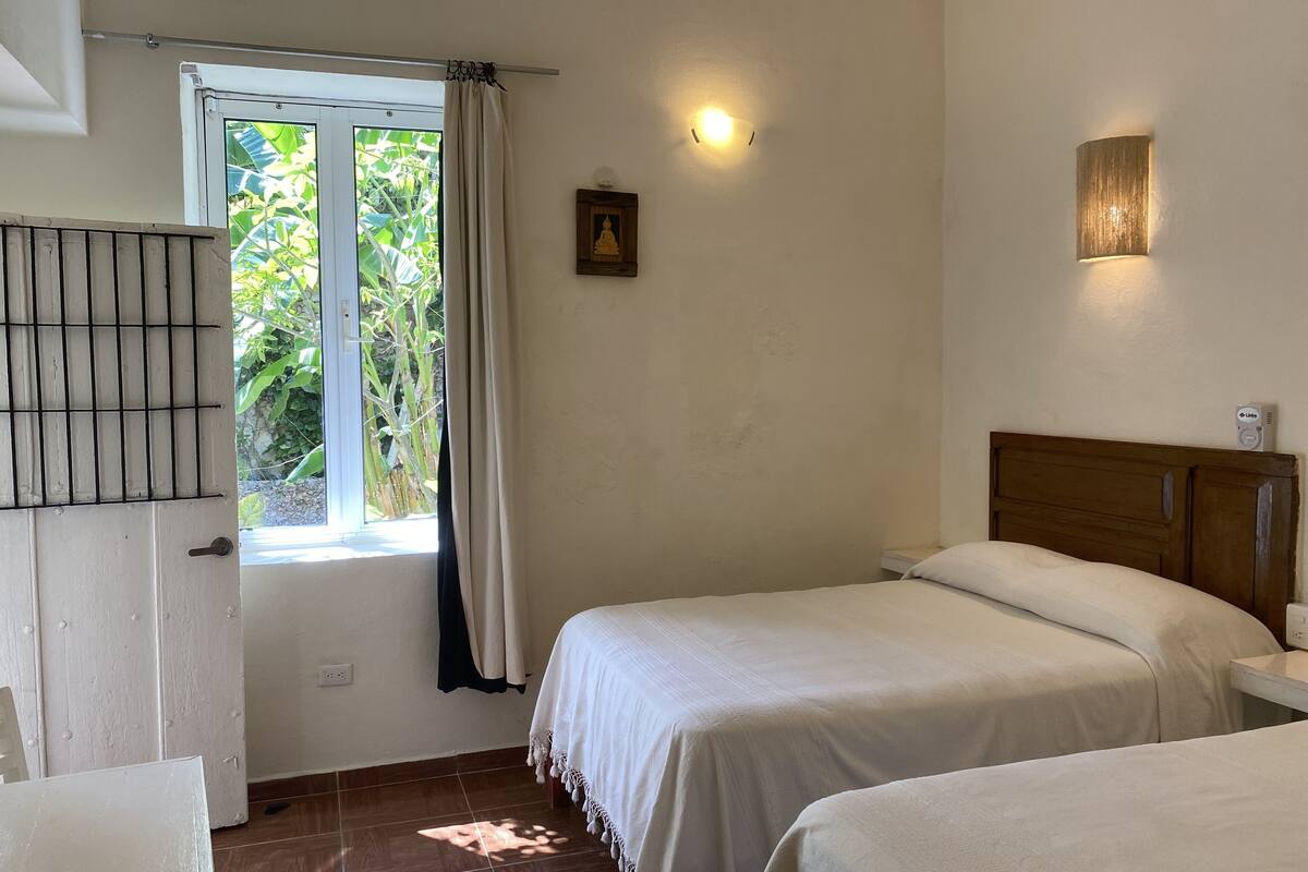 A bedroom with two beds, a window with curtains, a wall-mounted light, and a small Buddha statue.