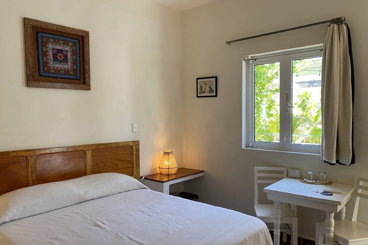 A bedroom with a bed, a bedside table with a lamp, a window with curtains, a small table with chairs, and a framed picture on the wall.