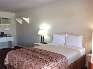 Ein Hotelzimmer mit einem Bett, einem Nachttisch mit Lampe, einer Wand-Klimaanlage und einem kleinen Schreibtisch mit Kaffeemaschine.