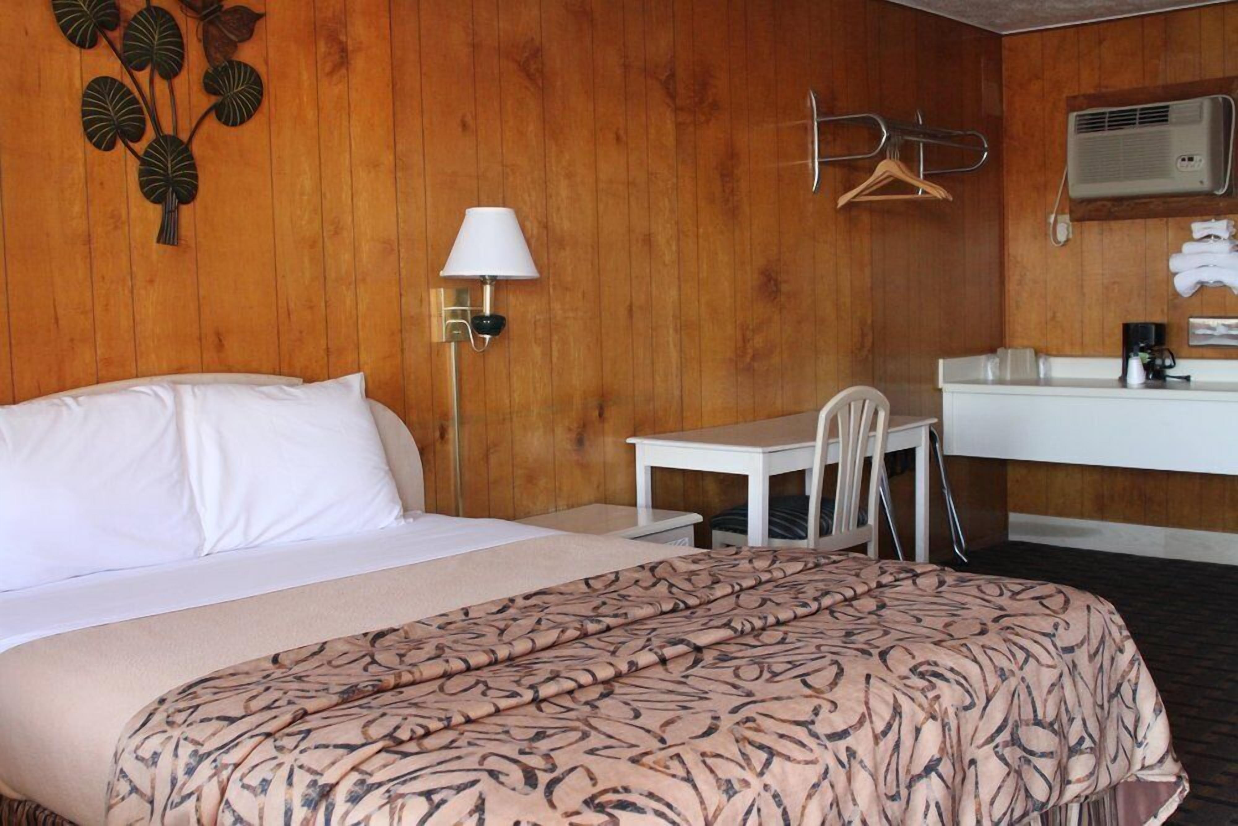 Ett hotellrum med en säng, ett skrivbord, en stol, en klädstång och ett badrum med en spegel och en handduksställning.