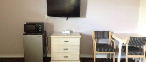 A hotel room with a flat-screen TV mounted on the wall, a small refrigerator with a microwave, a wooden chair with a striped cushion, and a white dresser with drawers.