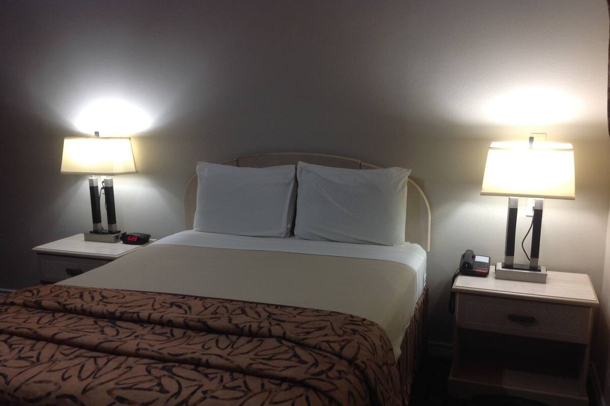 Quarto de hotel com uma cama, duas mesas de cabeceira, uma luminária em cada mesa e um relógio em uma delas.