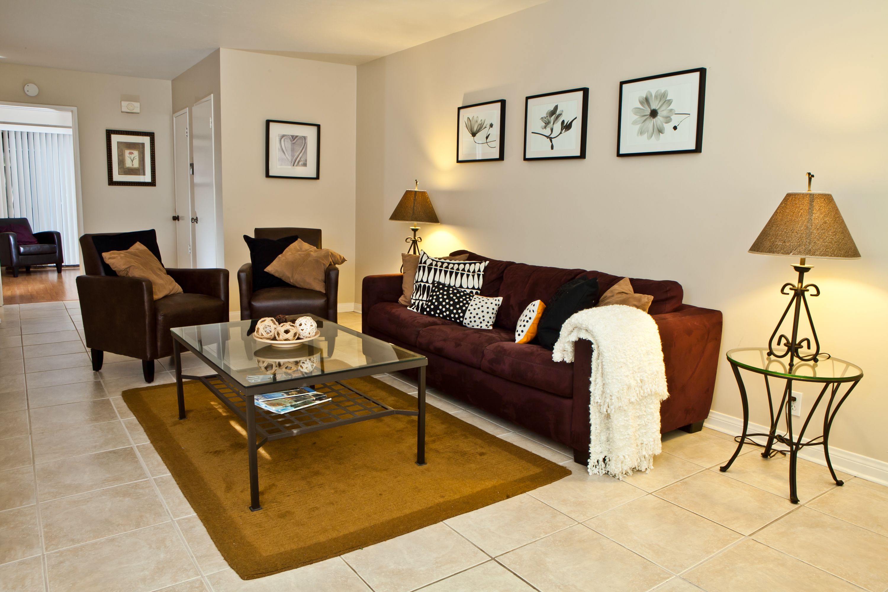 A living room with a glass coffee table, a brown sofa, and a red sofa. There are two lamps on side tables, and a brown rug on the floor.