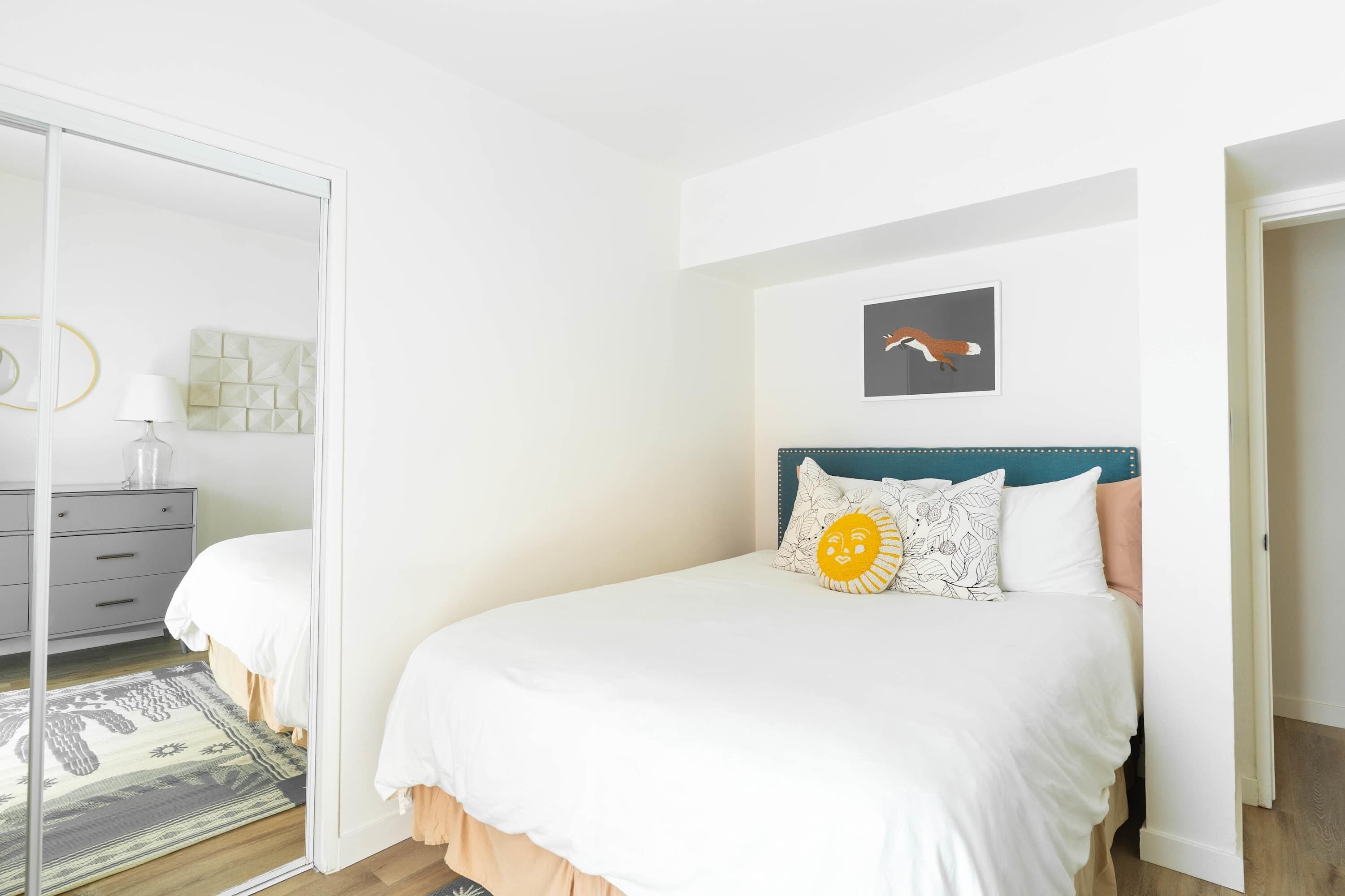Une chambre à coucher comprenant un lit, une table de chevet, une lampe, un tableau décoratif et une porte donnant sur une autre pièce.