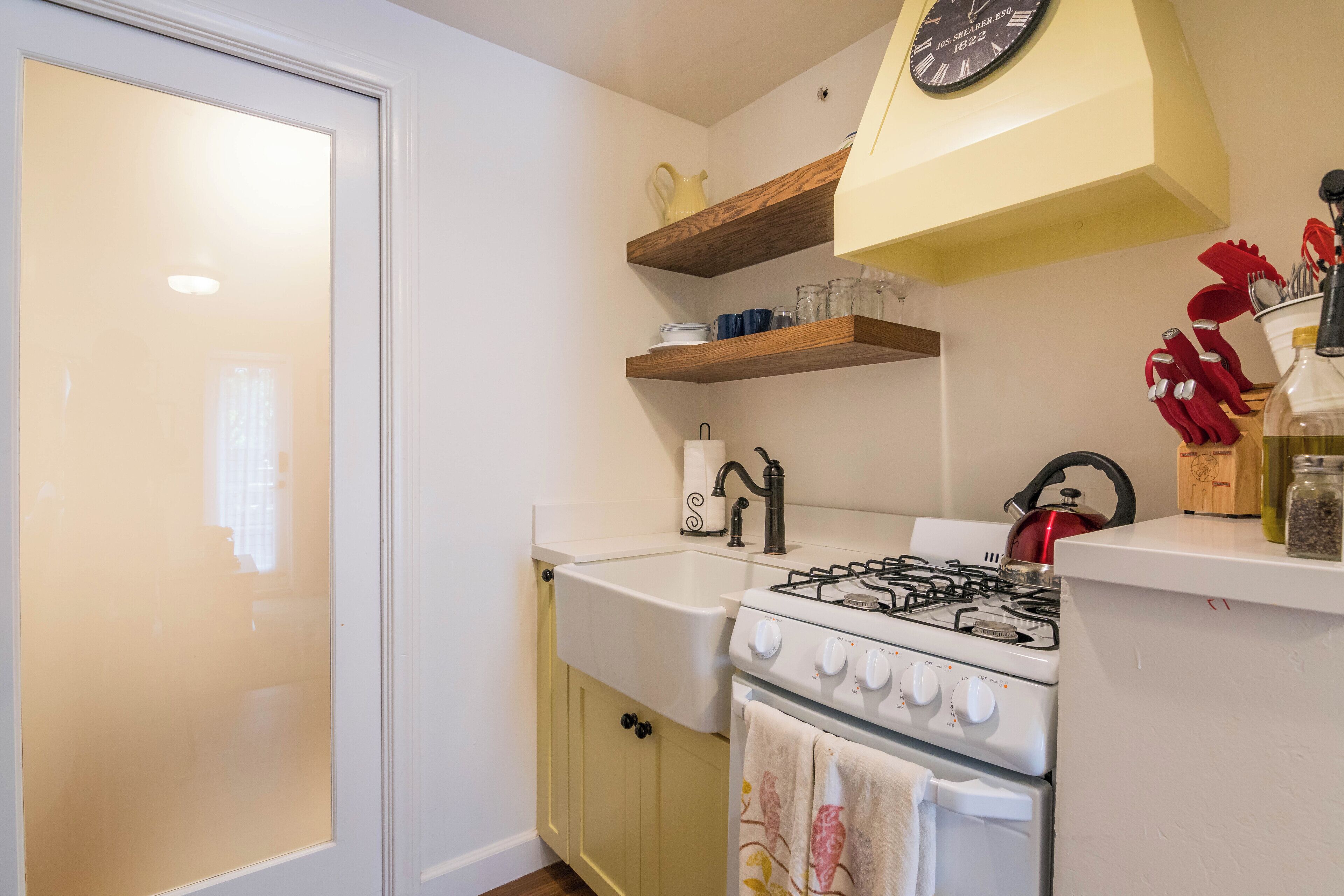 A kitchen with a white sink, stove, and oven, wooden shelves above, and a clock on the wall.