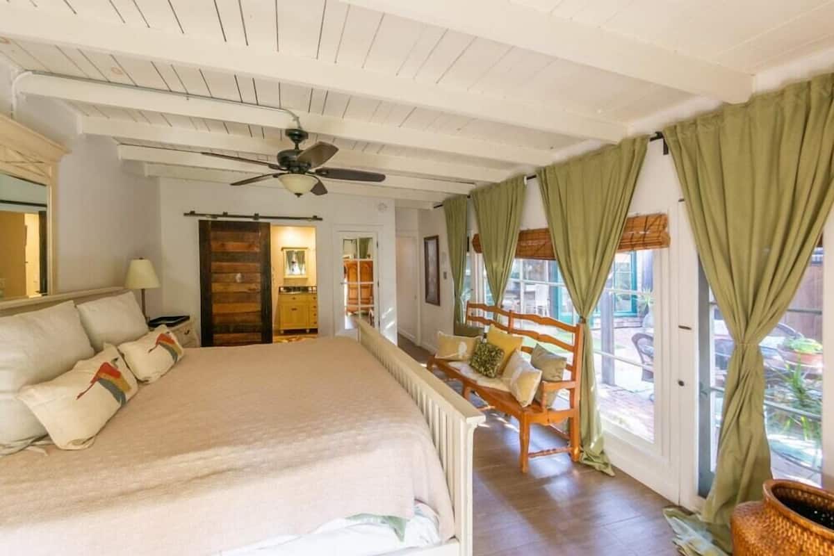 Un dormitorio con una cama grande, un ventilador de techo, una puerta de madera y vistas al exterior a través de un cristalero corredizo.