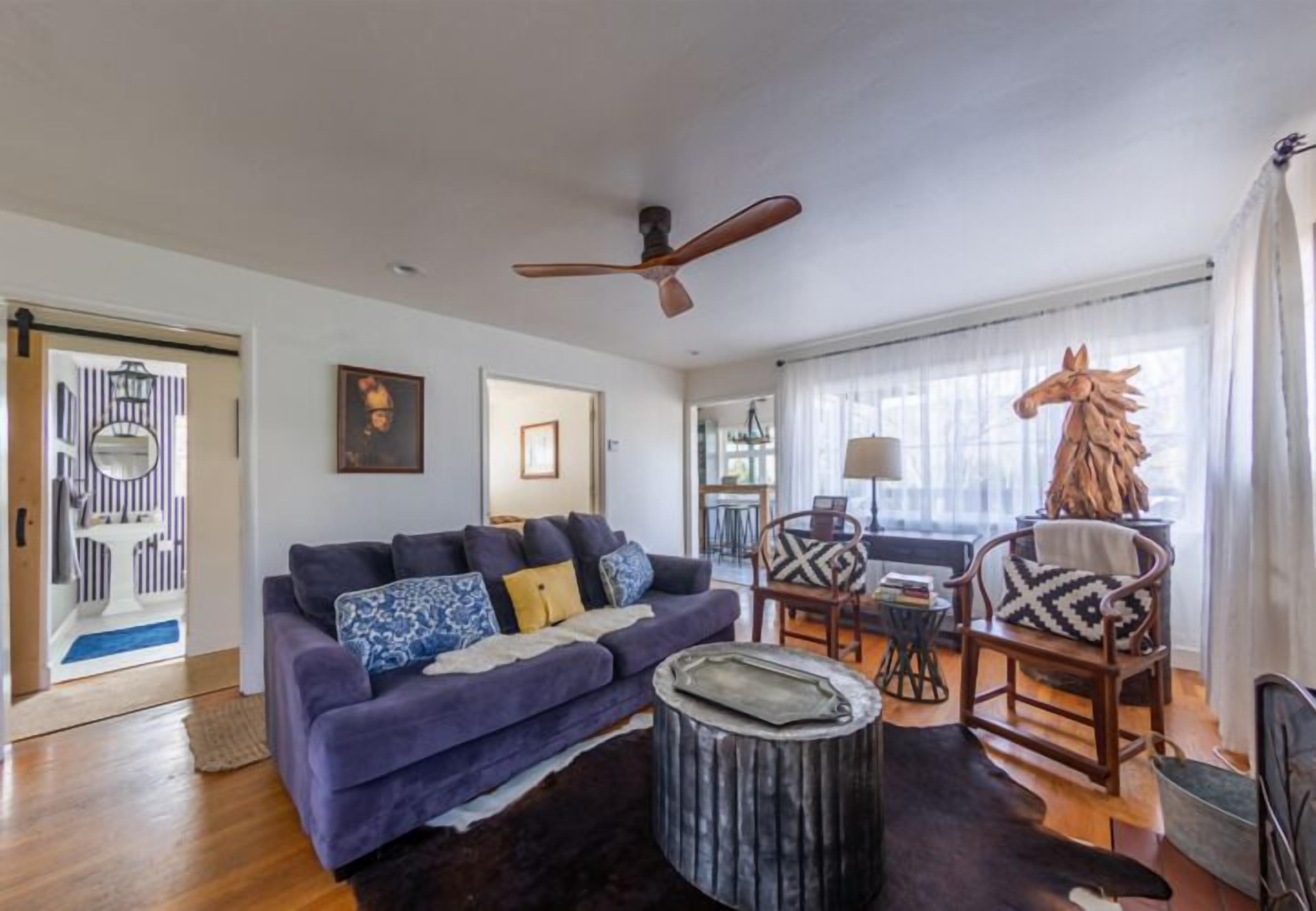 A living room with a purple sofa, wooden chairs, a round coffee table, a ceiling fan, and a large window with curtains.