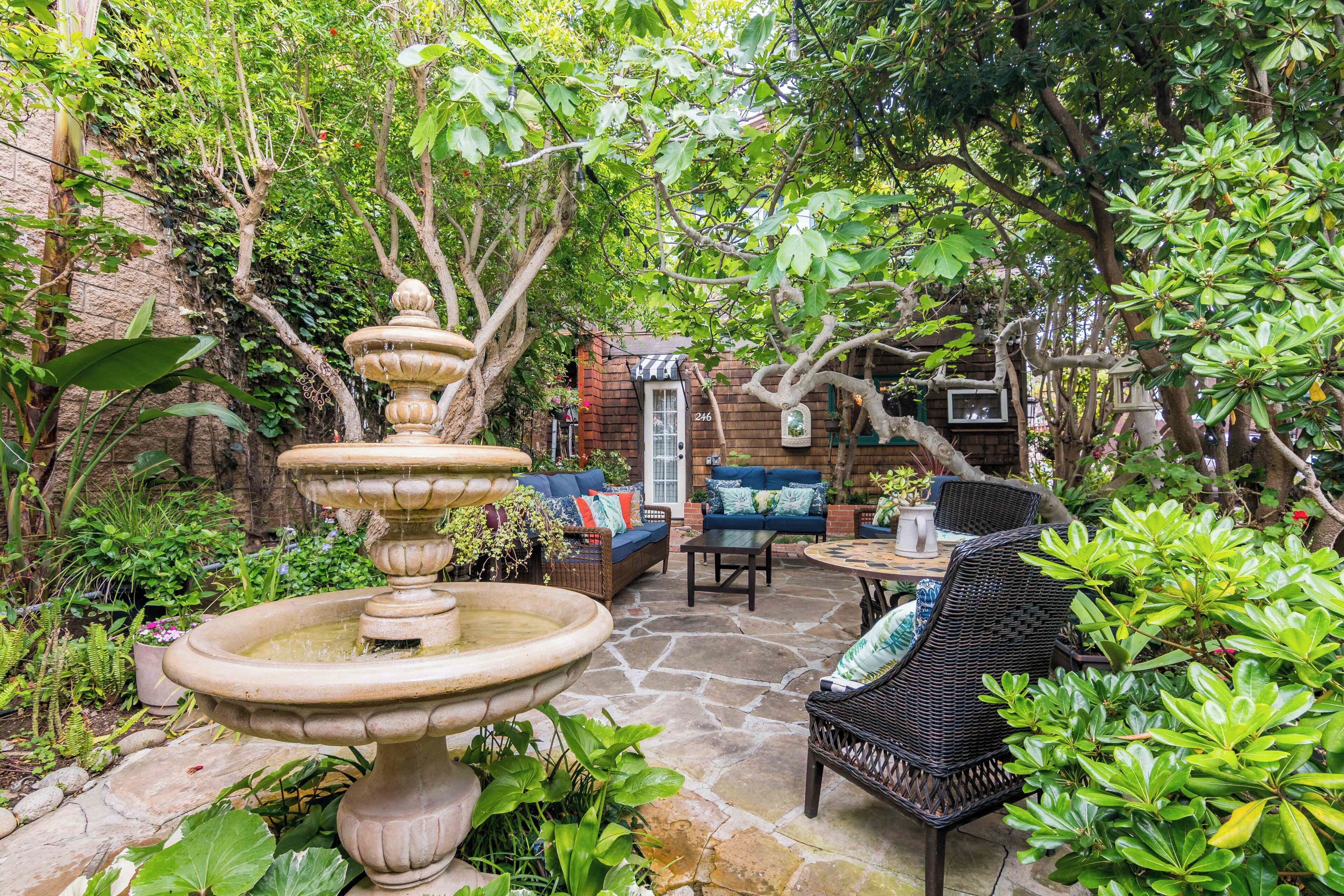 A stone patio with a tiered fountain, wicker furniture, and lush greenery.