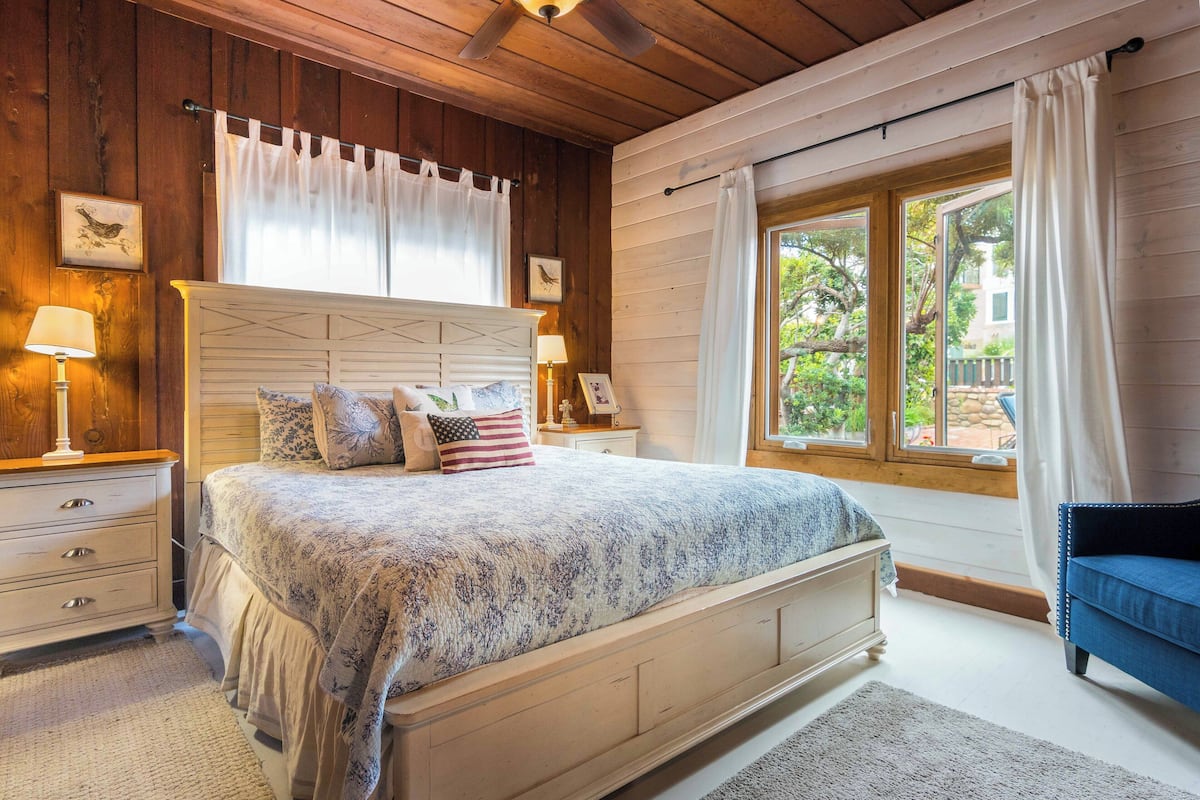 Un dormitorio con una cama grande, paredes de madera, una ventana con cortinas y una mesita de noche con lámpara.