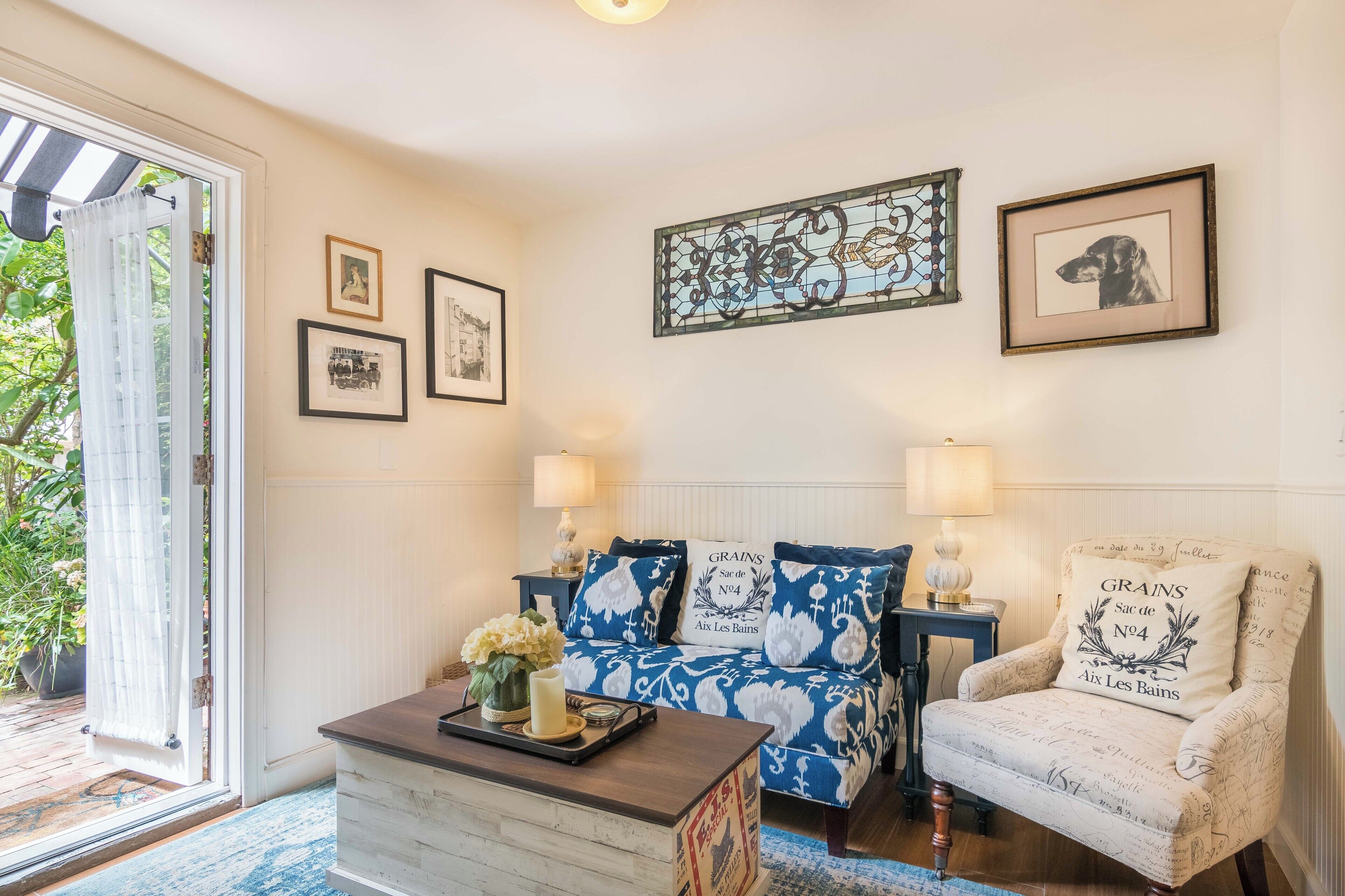 A cozy room with a sofa, armchair, and a coffee table. There are framed pictures on the wall and a window with curtains.